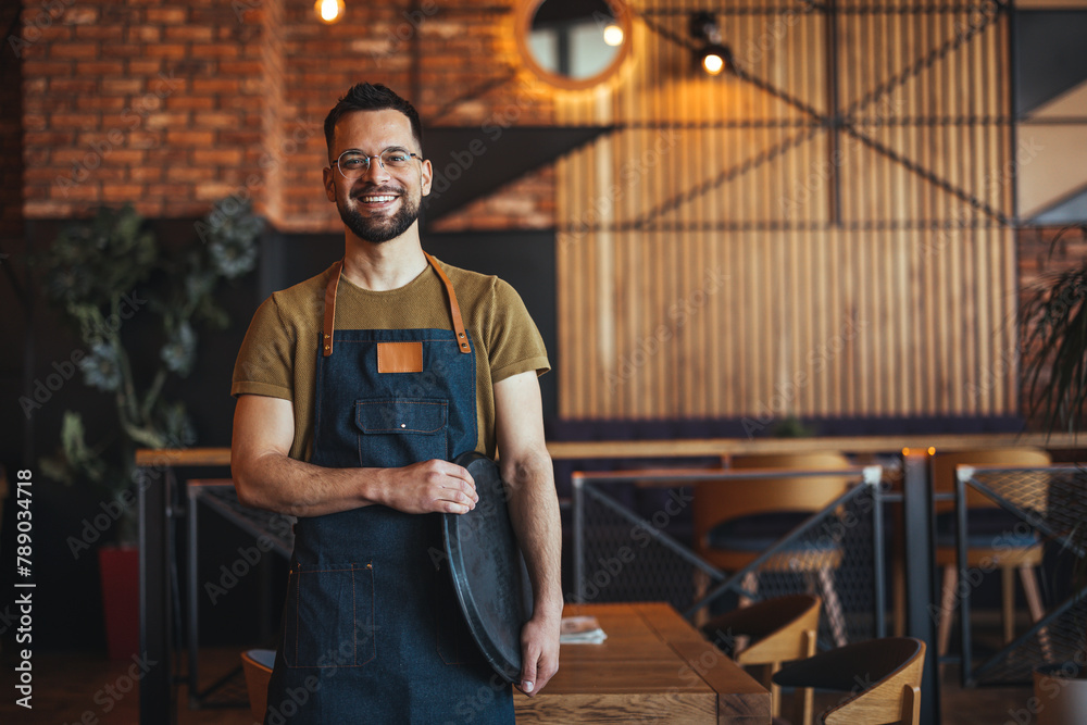 Obraz premium Waist up portrait of handsome waiter smiling cheerfully at camera standing in restaurant or cafe, copy space. Young waiter serving coffee in a cafe and looking at camera.