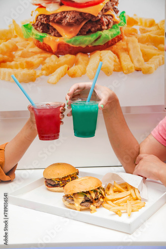 Hands of two women cheers holding lemonade slush drinks with burgers and fries on the table.