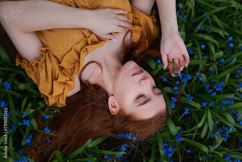 Young red haired woman in vintage dress laying on grass with blue flowers. Romantic top view portrait of woman with closed eyes