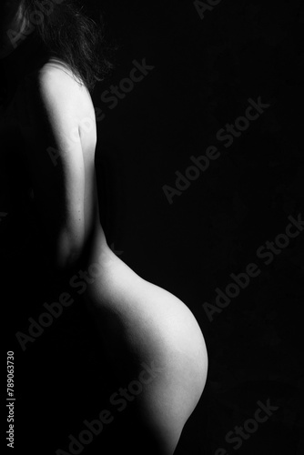Nude, erotic concept. Woman posing naked in studio. Model standing in shadow. Girls naked body silhouette illuminated with light. Image contains noise and motion blur. Black and white image