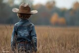 A young boy in a cowboy hat stands contemplatively in a vast, open field during autumn