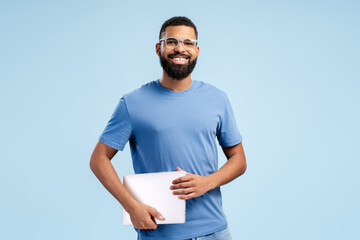 Happy African American in eyeglasses holding laptop while looking at camera, over blue background