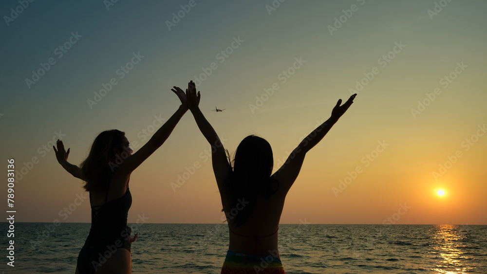 Two women are standing on the beach while happily watching an airplane pass above them.