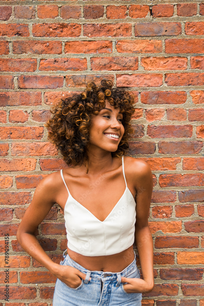 A smiling black woman with curly hair stands confidently in a white top and denim in front of a textured brick wall, exuding joy and style