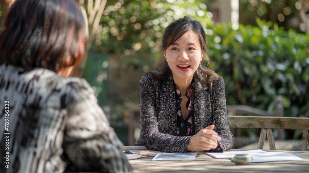 Relaxed outdoor meeting in a sunny park, with an Asian businesswoman sitting across from a client at a rustic wooden table, discussing plans with a smile, in a bright, realistic style. Relaxed outdoor meeting in a sunny park, with an Asian businesswoman sitting across from a client at a rustic wooden table, discussing plans with a smile, in a bright, realistic style.