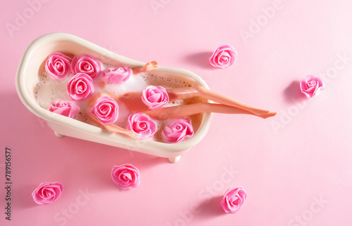Top view doll in bath with soap foam and pink flowers on a pink background.