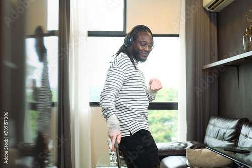 Carefree happy young African man listening to music in headphones, enjoying housework