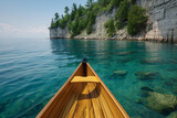 The bow of a wooden canoe navigates through the clear waters of a serene lake, flanked by rugged cliffs and lush greenery under a vibrant blue sky.