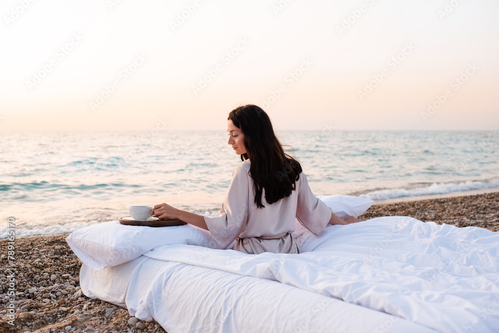 Beautiful young woman resting in bed over sea having morning coffee outdoor. Summer season.