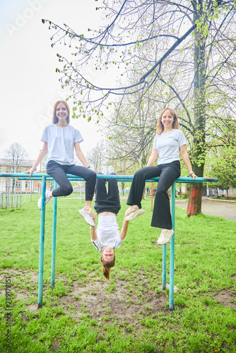 Three girls of high school age in physical education uniforms are sitting on a horizontal bar. One is hanging upside down.