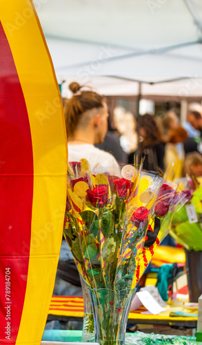 Bouquet of roses decorated with ears of wheat and bows of the flag of Catalonia, at a flower and book stall in a traditional holiday market with people walking on Sant Jordi day.