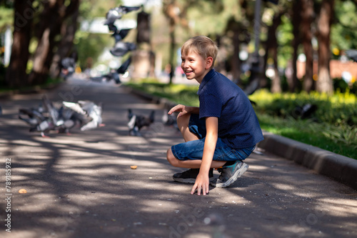 A boy in a blue T-shirt sits on the asphalt in the park and looks at the pigeons.
