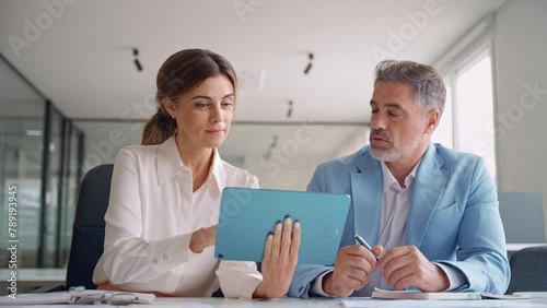Smiling mature female executive manager holding digital tablet consulting client discussing tech services at corporate meeting. Two happy professional business colleagues working sitting in office.