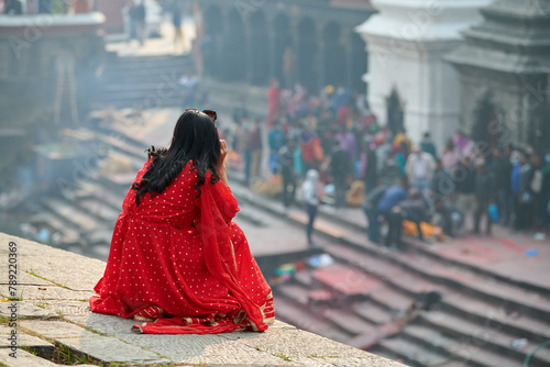 Back view of woman in red sari watching mournful cremation ceremony at Pashupatinath Temple complex, Nepal, view from Pandra Shivalaya site, symbolizing cyclical nature of life and death photo