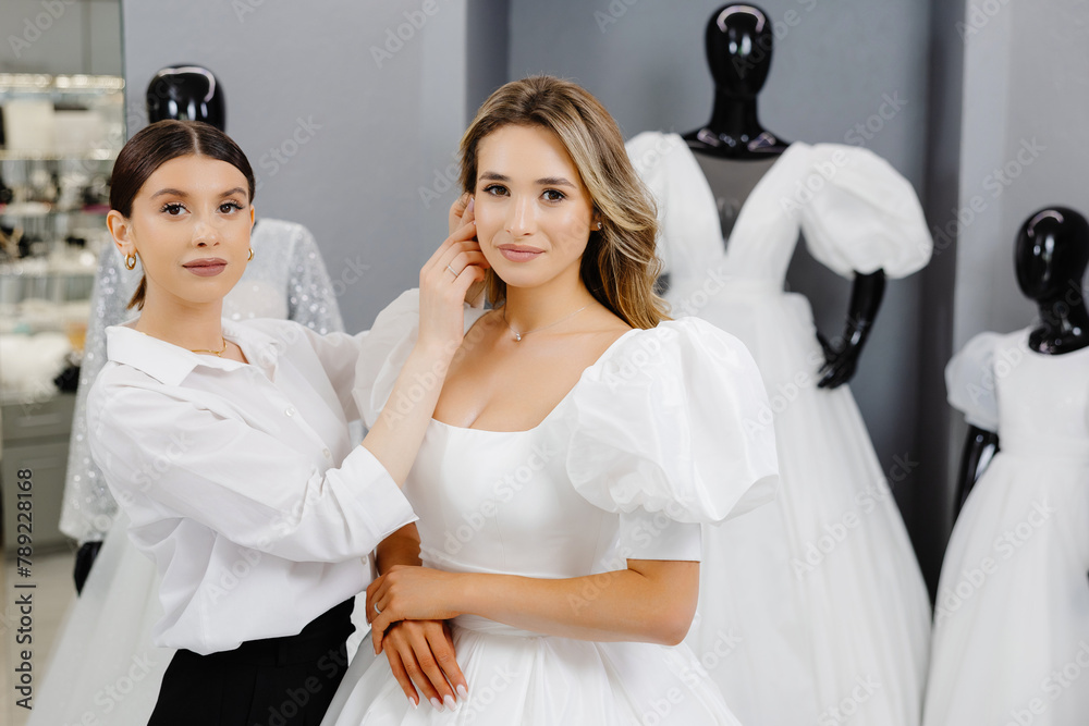 The bride-to-be came to a wedding dress salon to buy a dress. A beautiful woman chooses a wedding dress in the salon, and an assistant helps the bride put on an earring The bride-to-be came to a wedding dress salon to buy a dress. A beautiful woman chooses a wedding dress in the salon, and an assistant helps the bride put on an earring