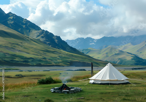 A big white camping tent with camping gears in a green grass with mountains and blue sky with white clouds in the background. Taken during golden hour