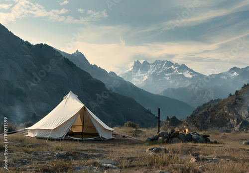 A big white camping tent with camping gears in a green grass with mountains and blue sky with white clouds in the background. Taken during golden hour