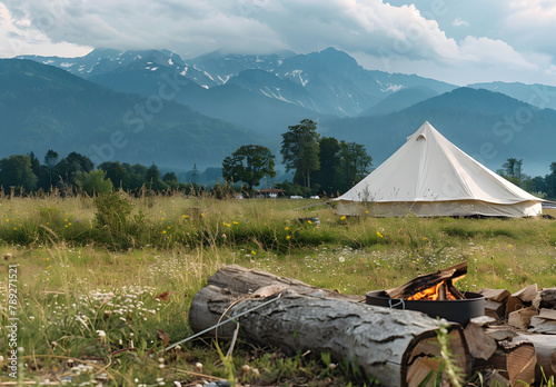 A big white camping tent with camping gears in a green grass with mountains and blue sky with white clouds in the background. Taken during golden hour