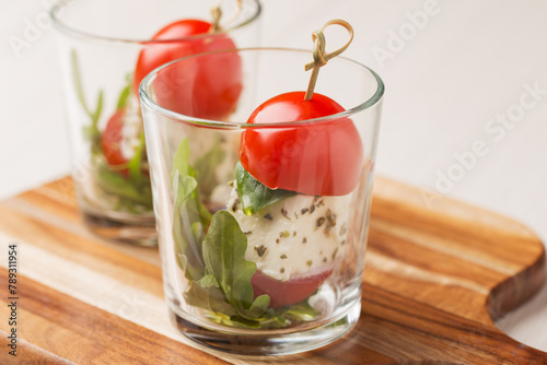 Canapes caprese on skewers with tomatoes, mozzarella, basil and olive oil in glasses on a light wooden table. Mediterranean cuisine. Selective focus.
