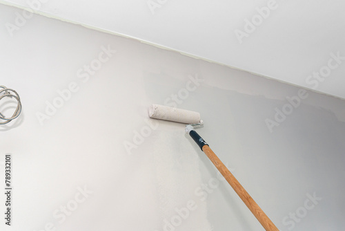 Painter paints a room in beige color using a paint roller on an extended stick at home.