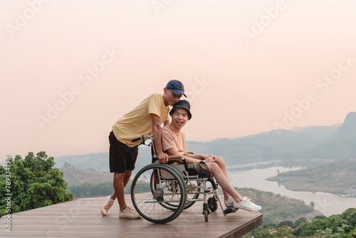 A parent, caregiver or volunteer and a confident young man on wheelchair with lighting sunset, Happy family time and learning on the natural park,Travel on vacation with family, Mental health concept.