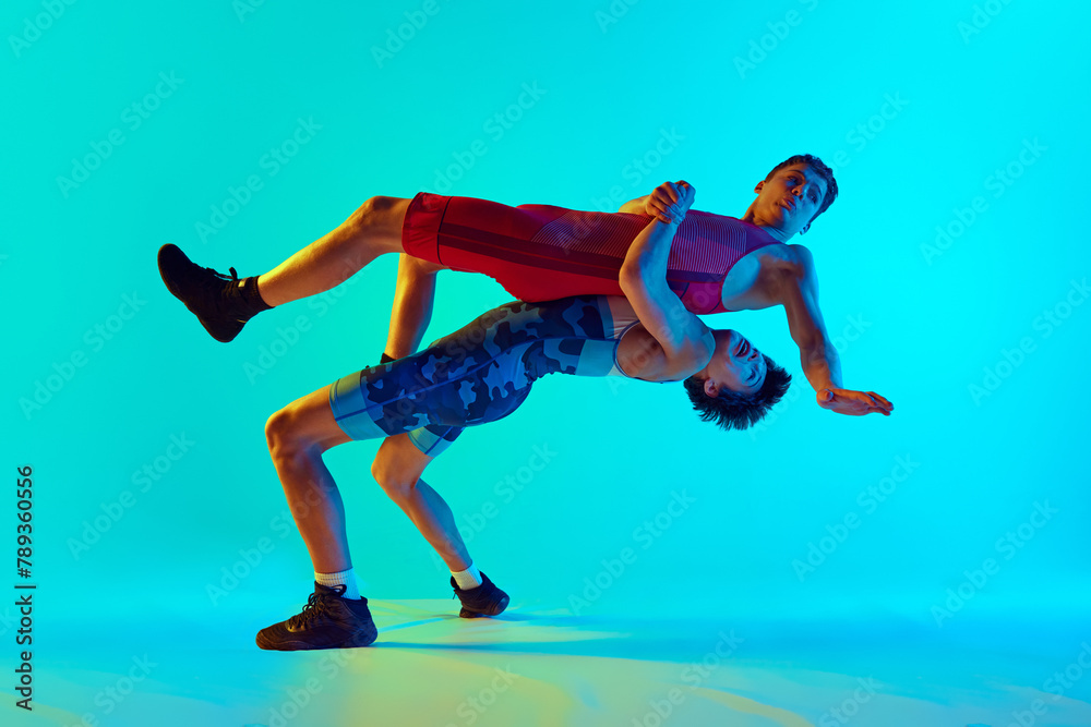 Young men, athletes demonstrate focus and strength as they engage in competitive wrestling match against blue background in neon light. Concept of combat sport, martial arts, competition, tournament