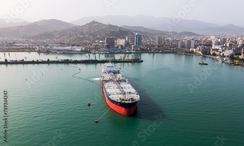 Aerial view of tanker ship in clear turquoise waters near coastal cityscape. © DedMityay