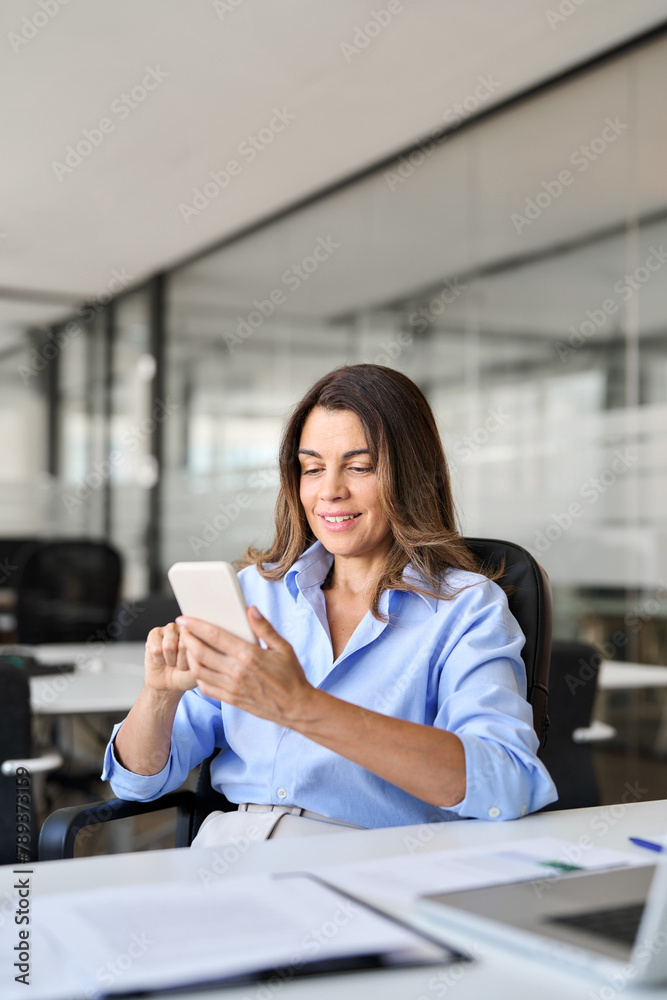 © insta_photos - Smiling mature business woman executive using mobile phone working in office. Happy middle aged businesswoman holding smartphone, professional female executive looking at cellphone at work. Vertical.
