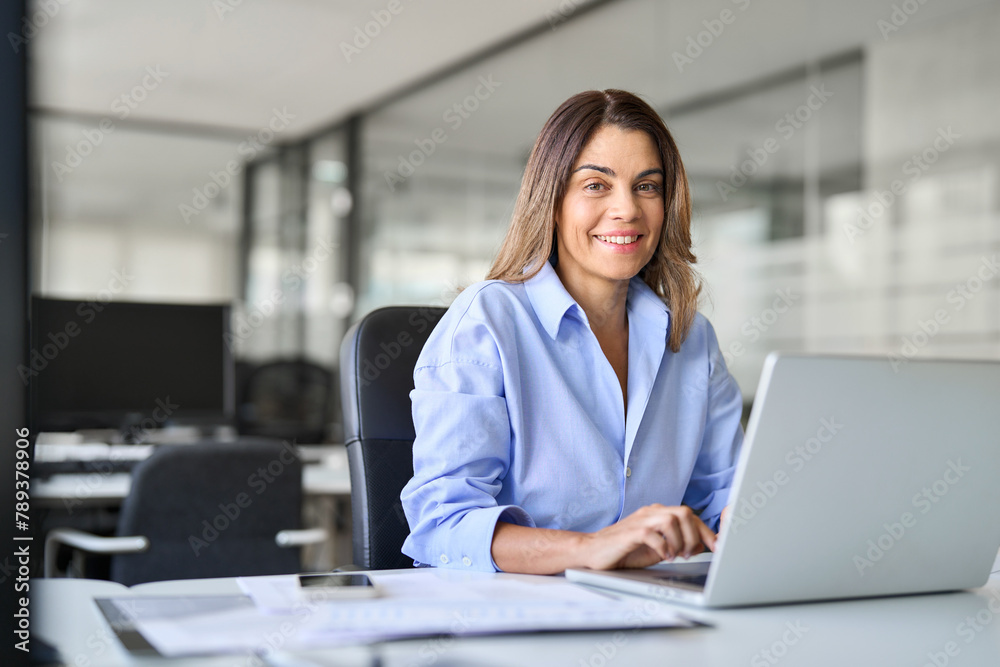 Happy smiling mature middle aged professional business woman bank worker or hr manager, corporate executive or lawyer attorney looking at camera at work working on laptop computer in office, portrait. Happy smiling mature middle aged professional business woman bank worker or hr manager, corporate executive or lawyer attorney looking at camera at work working on laptop computer in office, portrait.