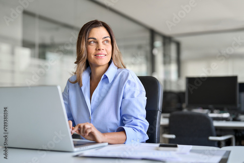 Happy mature middle aged professional business woman bank worker or hr manager, corporate executive looking away at work working on laptop computer in office thinking of business technology future.