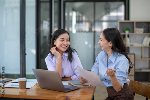 Two smiling businesswomen collaborating in a modern office environment, reviewing a document and using a laptop.