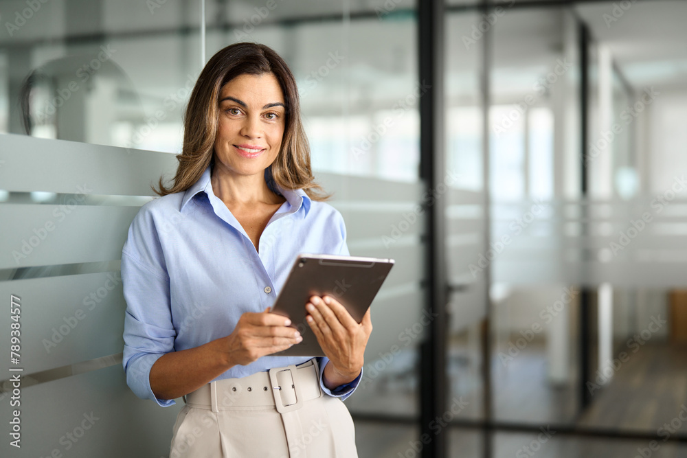 Obraz premium Smiling mature business woman executive, happy middle aged businesswoman entrepreneur, 40 years old company hr holding digital tablet looking at camera standing in office at work. Portrait.