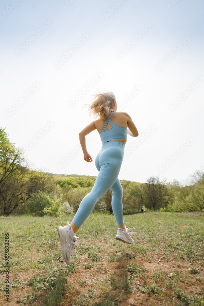 Bottom view of an athletic girl jogging in the park. Beautiful blonde Caucasian woman in blue tight tracksuit. Blonde girl at an outdoor training session