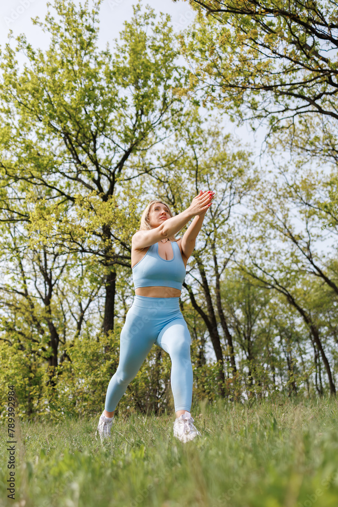 Girl athlete training in the park stretched her arms in front of her and performs lunges forward. Beautiful blonde Caucasian woman in blue tight tracksuit. Blonde girl at an outdoor training session