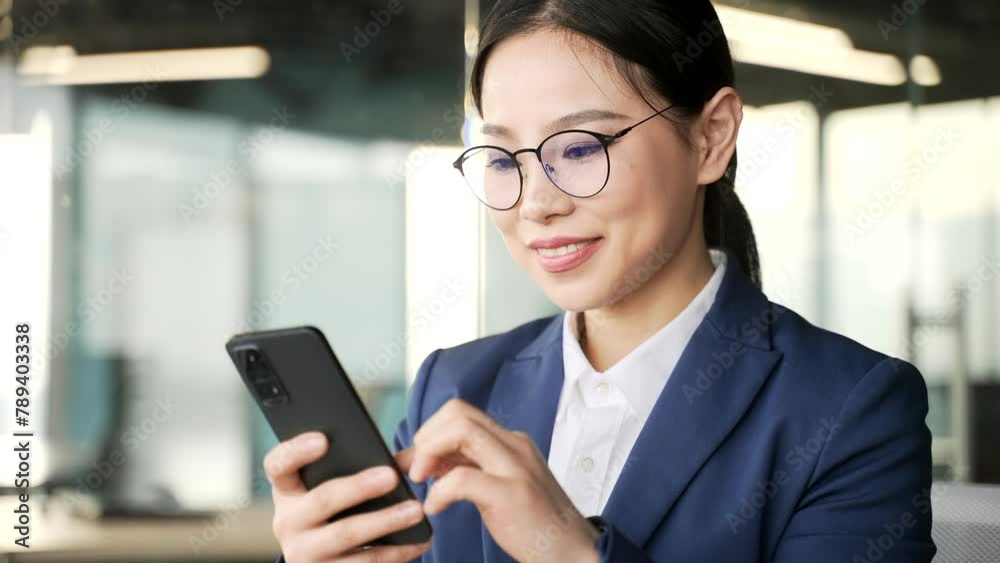 Asian businesswoman is using phone sitting at workplace in business office. Woman employee in formal suit works on mobile application, reading typing a message, chatting or browsing online. Close up Asian businesswoman is using phone sitting at workplace in business office. Woman employee in formal suit works on mobile application, reading typing a message, chatting or browsing online. Close up