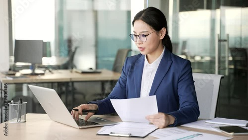 Busy young asian businesswoman female accountant doing paperwork using laptop computer sitting at workplace in business office. Woman financier in glasses, making financial report or writing tax form photo