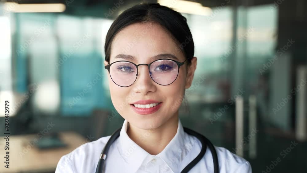 Portrait of smiling asian female pediatrician in white coat wearing glasses looking at camera in modern hospital clinic. Happy woman medical worker doctor poses standing in the office. Close up Portrait of smiling asian female pediatrician in white coat wearing glasses looking at camera in modern hospital clinic. Happy woman medical worker doctor poses standing in the office. Close up