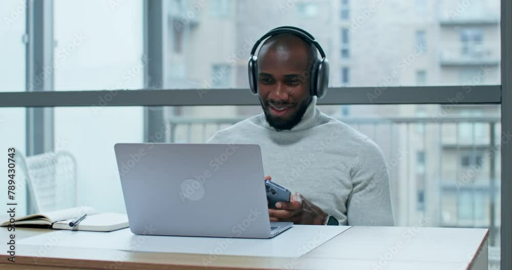 A man wearing headphones uses a mobile phone sitting at a table indoors. An open laptop stands in front of him A man wearing headphones uses a mobile phone sitting at a table indoors. An open laptop stands in front of him