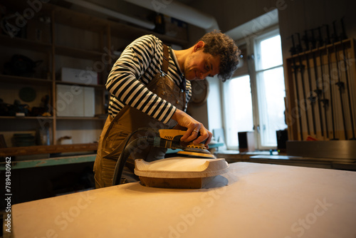 Sanding wood with orbital sander at workshop. Focused man carpenter polishes wooden seat of a future chair with electric sander. Carpentry workshop. Furniture production