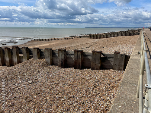 View of Bexhill on Sea seafront with groins and railing by promenade looking towards iconic Beach Head headland on a sunny blue sky day