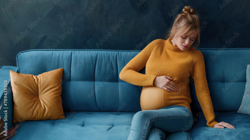 Obraz premium Pregnant young woman holding her belly, sitting on a sofa in the living room. Mother in pregnancy, abdomen cramp pain, prenatal lady discomfort, expecting a baby, lady trimester, copy space