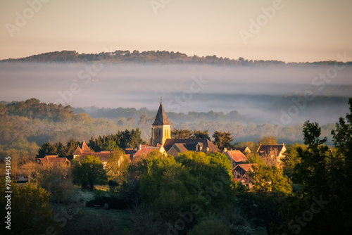 Sunshine on the village and church of Nabirat in the Dordogne region of France with early morning mist over the distant woodland