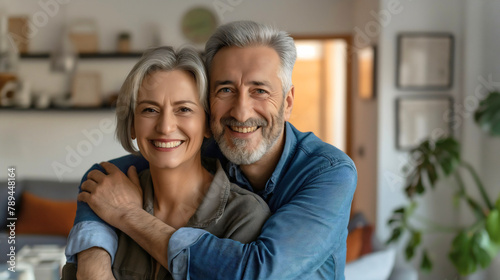 Middle aged man and woman, happy married couple love relationship, standing hugged in living room interior, smiling at camera. Mature husband wife home lifestyle indoors, embracing togetherness