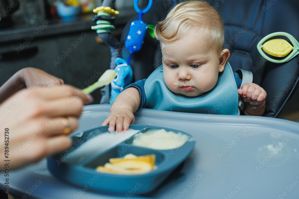 The first feeding of the baby is pureed complementary food. Baby food with a spoon. A mother feeds puree to her 6-month-old son. Parents feed their baby with food on the bed The first feeding of the baby is pureed complementary food. Baby food with a spoon. A mother feeds puree to her 6-month-old son. Parents feed their baby with food on the bed