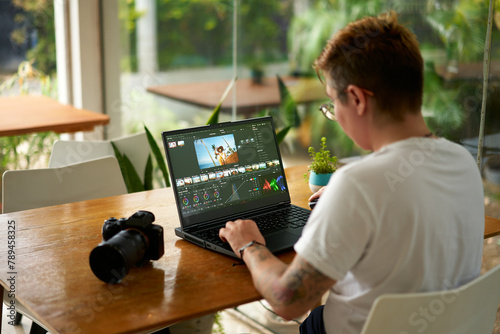 Fototapeta Naklejka Na Ścianę i Meble -  Pro videographer edits wildlife footage on laptop in a bright office. Color grading process by creative with camera gear beside. Concentrated freelancer tweaks travel video in a workspace.