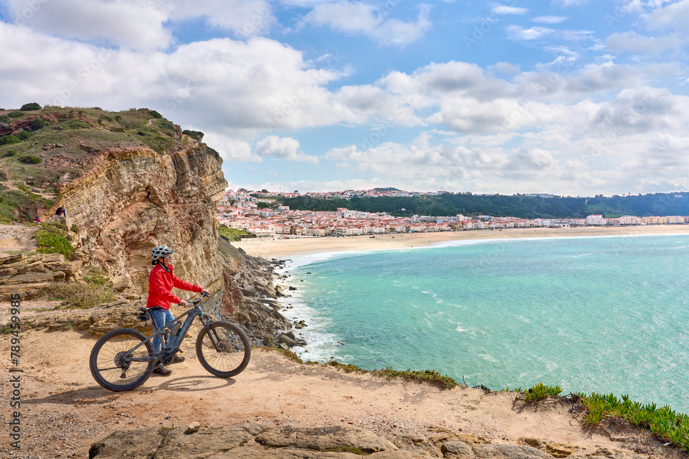 nice senior woman riding her electric mountain bike on the rocky cliffs above the city of Nazare at the western atlantic coast of Portugal
