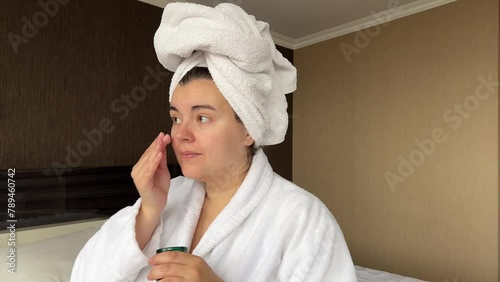 Horizontal 4K video. Young adult woman in white robe, towel on head applies natural moisturizer to face in hotel room. Skin protection during cold season. Cosmetic procedure in spa salon. Rest, relax