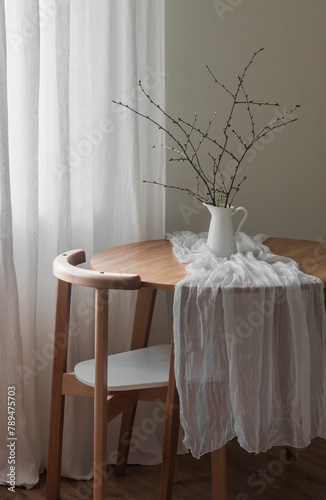 The simple interior of the living room - a round wooden table with a jug with branches and a chair. Minimalism style