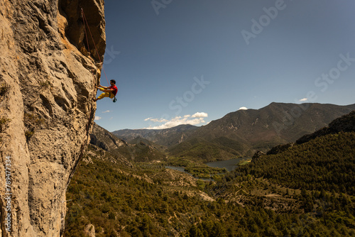 A man is climbing a rock face with a yellow shirt. The mountain range in the background is lush and green