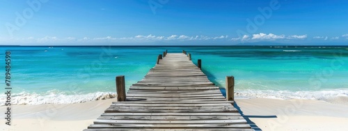 A white sand beach with a blue sky has a wooden pier that leads to the ocean.